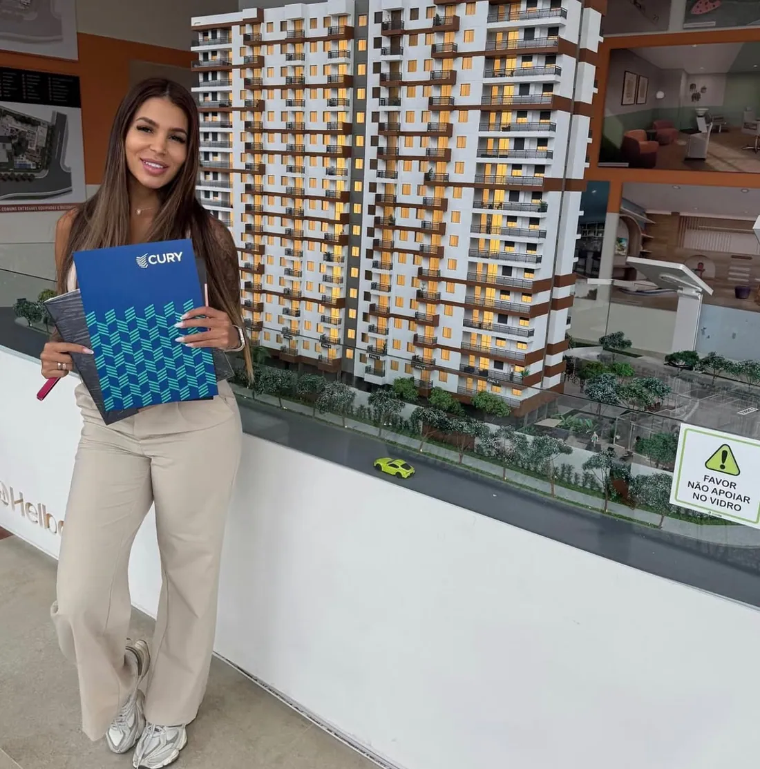 Luana segurando a pasta do contrato em frente à maquete do prédio